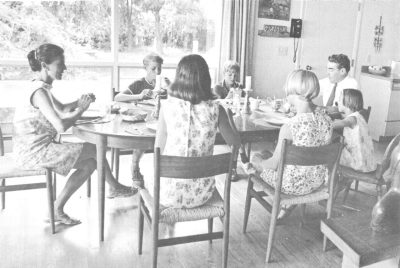 A black and white photo from 1969 shows the Carter family sitting around the dinner table eating. Mom and dad sit at each end of the table, while two boys sit facing the camera on one side of the table and three girls sit with their backs to the camera on the other side of the table. The room is bright and sits in front of a large window.