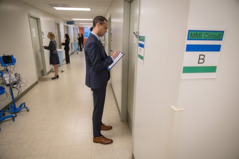 An individual in a business suit stands outside a doorway in a hall labeled MMI Circuit B writing on a paper on a clipboard.