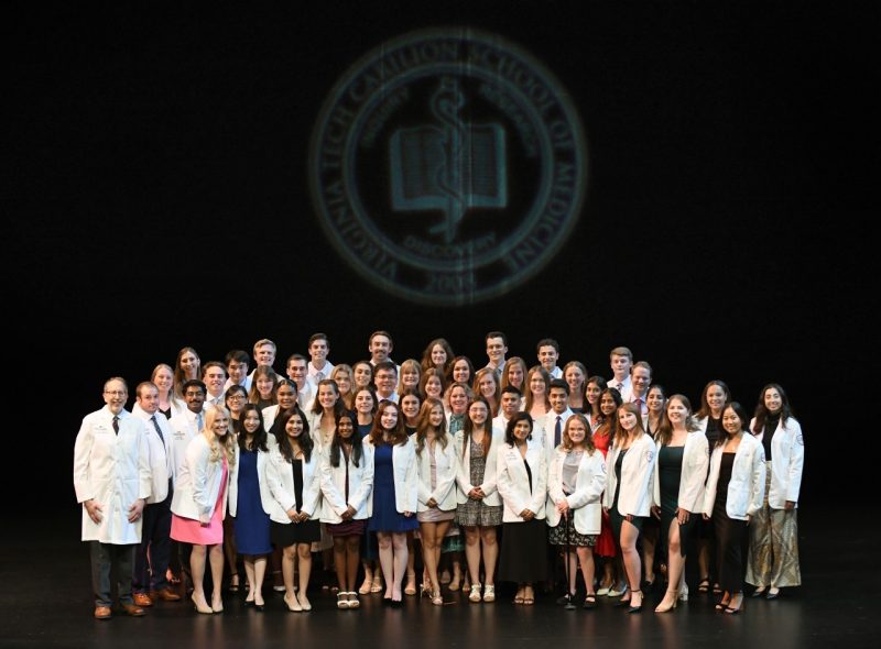 A group of diverse students wearing their physician white coats stand on a stage under the Virginia Tech Carilion School of Medicine seal projected on a wall.