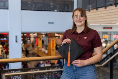 Haley Rindfleisch holds a graduation cap and tassel in her hands. She is wearing a maroon Virginia Tech shirt and blue jeans. She's standing against a stair railing in Hitt Hall.