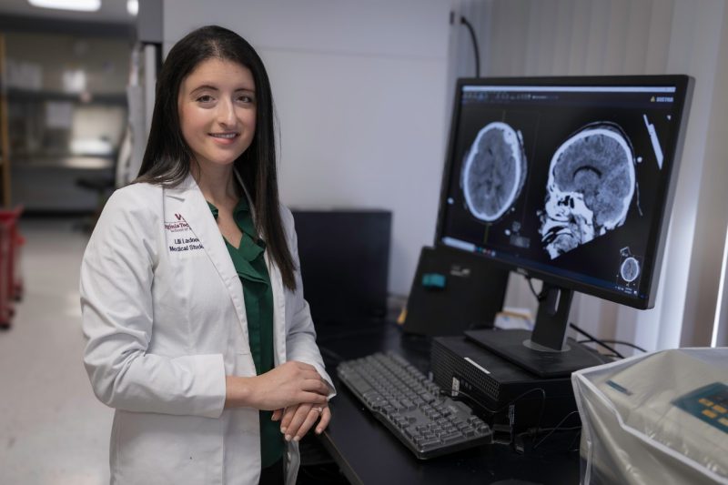 A medical student in a white coat stands in front of a computer screen with scans of brains.