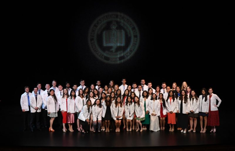 A large group of medical school students dressed in their white coats stand on stage underneath the school seal. 