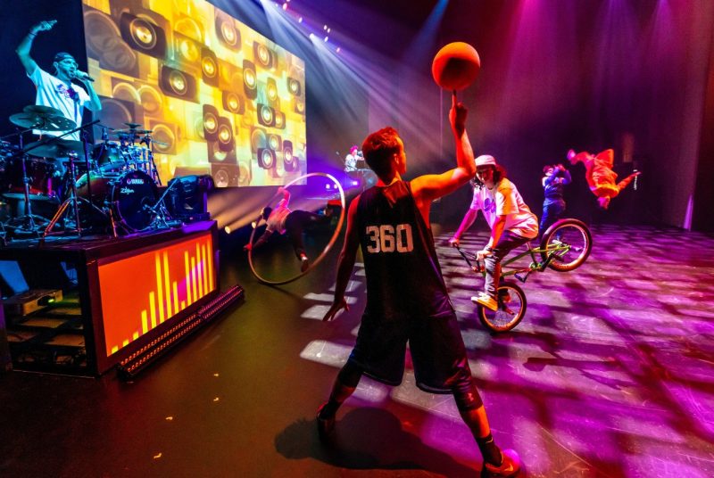 Performers from 360 ALLSTARS fill the stage with movement and color under vivid lights. A basketball player spins a ball on his finger while another rides a BMX bike on one wheel. Behind them, a drummer and DJ perform in front of a large, colorful video projection as acrobats flip and spin across the stage.