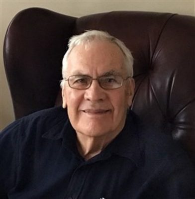 A man with gray hair and glasses is wearing a dark blue collared shirt. He is seated in a leather chair, smiling. 