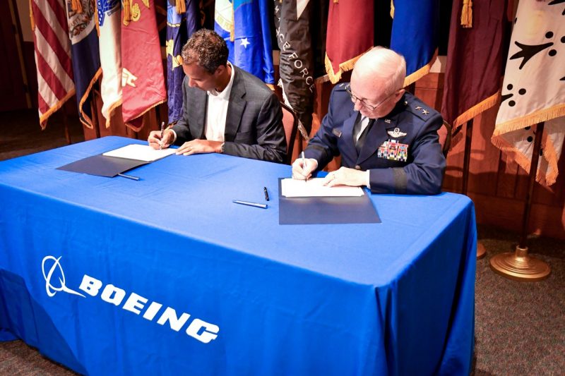 Marc Allen and Major General Fullhart sit at a clothed table with flags stationed in the background. Both are looking down and signing the memorandum.
