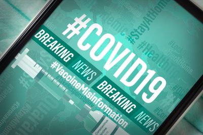 A phone screen reads "#COVID19 Vaccine Misinformation" along with two "Breaking News" tickers scrolling beneath it. Hashtags such as #StayAtHome and #Pandemic are scattered in the background